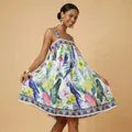 Woman smiling in a flowy tropical print sundress with blue parrots, yellow flowers, and green leaves, wearing gold hoop earrings and a gold cuff bracelet
