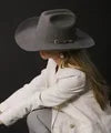 Woman wearing a grey felt cattleman cowboy hat with silver buckle band and white blazer, head tilted down, dark studio background
