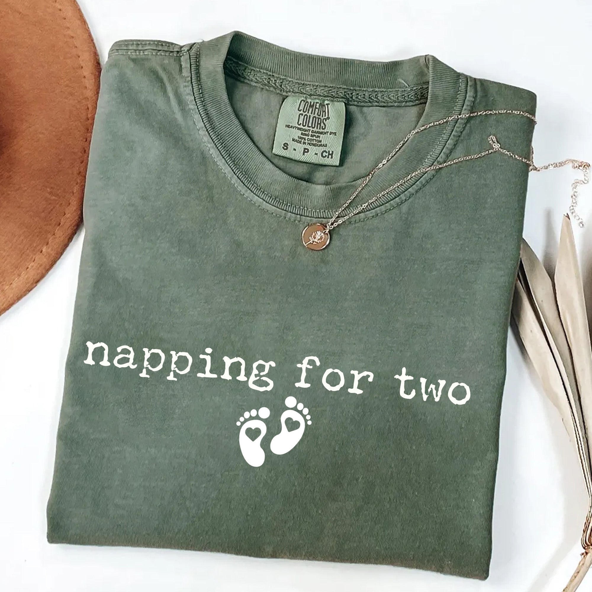 Folded green shirt with 'napping for two' text and footprints on a white background