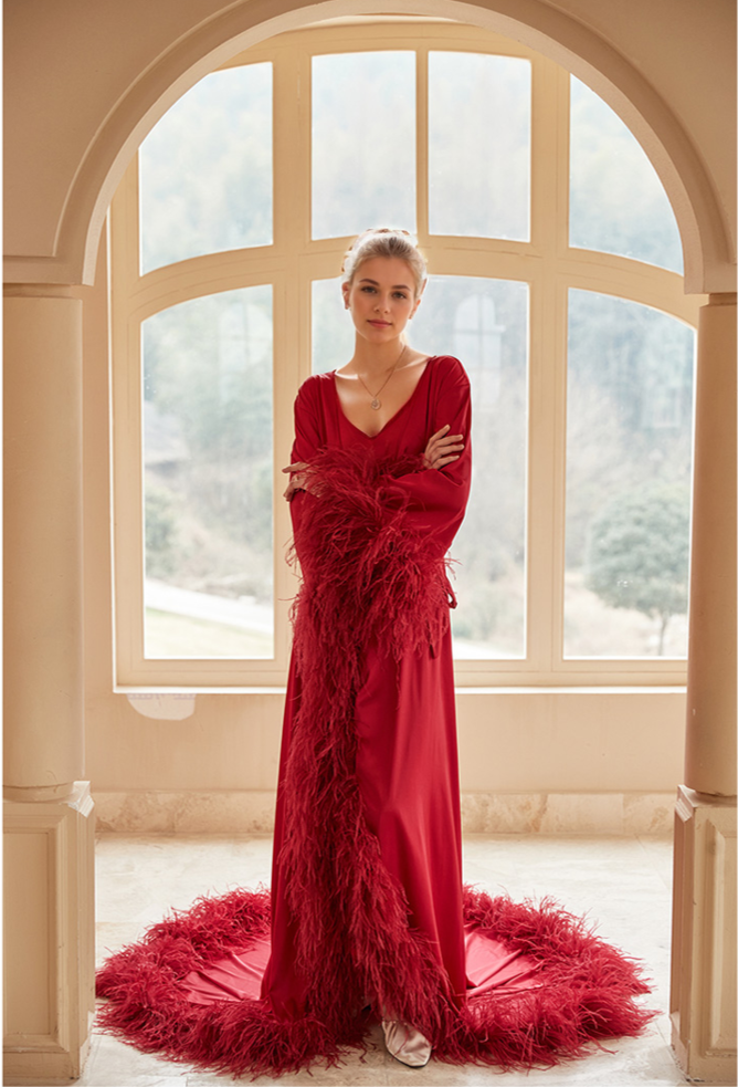 Woman in a red evening gown with feather details standing in front of large windows.