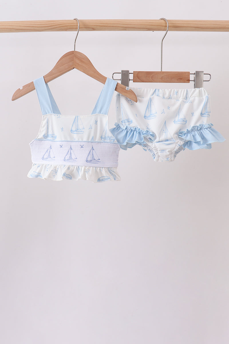 A two-piece baby swimsuit set hanging on wooden hangers from a wooden rod. The set includes a light blue and white ruffled tankini top with a sailboat print, and matching ruffled bloomers.
