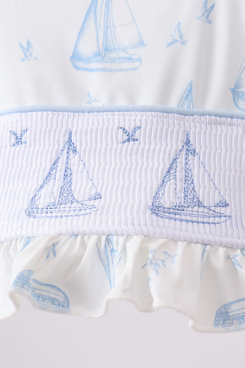 Close-up of a white ruffled garment with a smocked band featuring blue sailboat and bird motifs, plus a light blue sailboat print on the fabric.