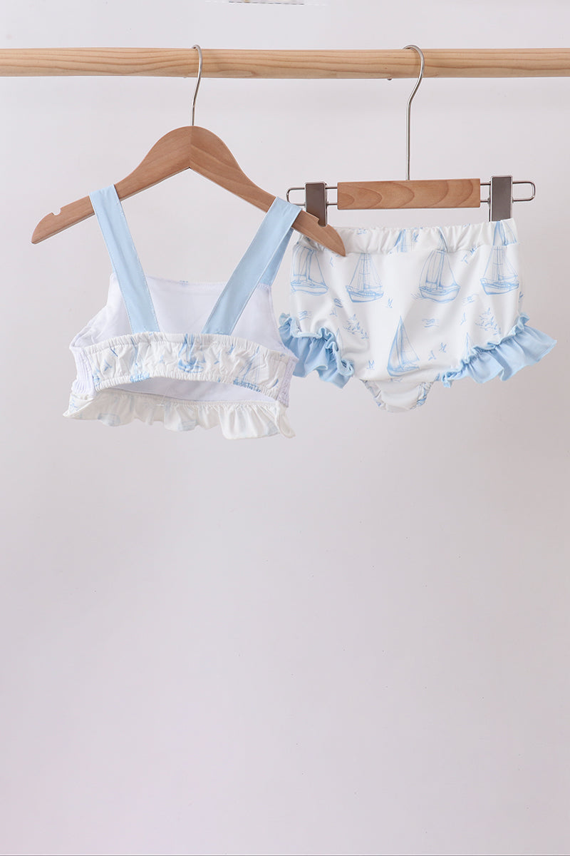 A baby two-piece swimsuit or romper set hanging on wooden hangers from a wooden clothes rack. The set includes a white tankini-style top with light blue straps and ruffled trim, and matching white bloomers with light blue ruffled trim and a subtle pattern.