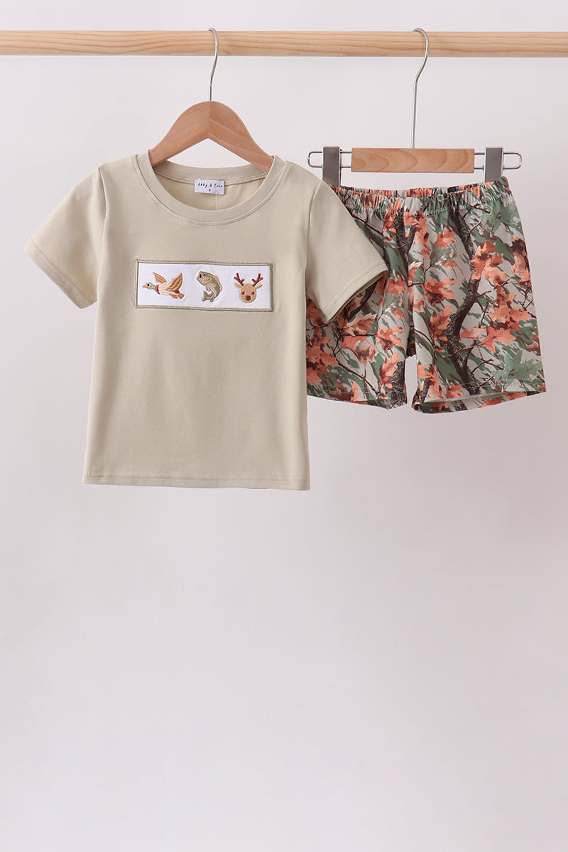A children's two-piece clothing set hanging on wooden hangers from a wooden rod. The set includes a short-sleeve t-shirt in a light beige color with a rectangular chest graphic showing three small animal illustrations, and matching shorts with a dense orange, green, and brown floral print.