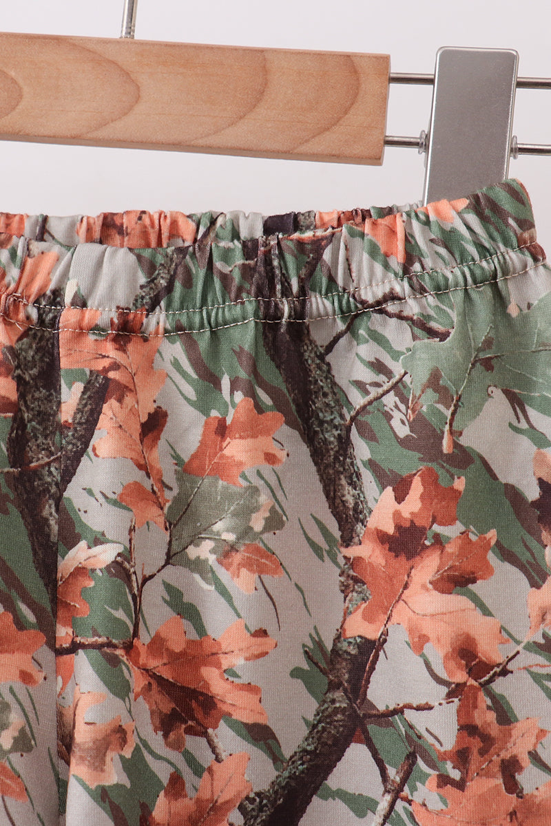 Close-up of a pair of camouflage-print pants hanging on a wooden hanger. The fabric features an autumn leaf and tree branch pattern in shades of green, brown, and terracotta orange on a light gray base.