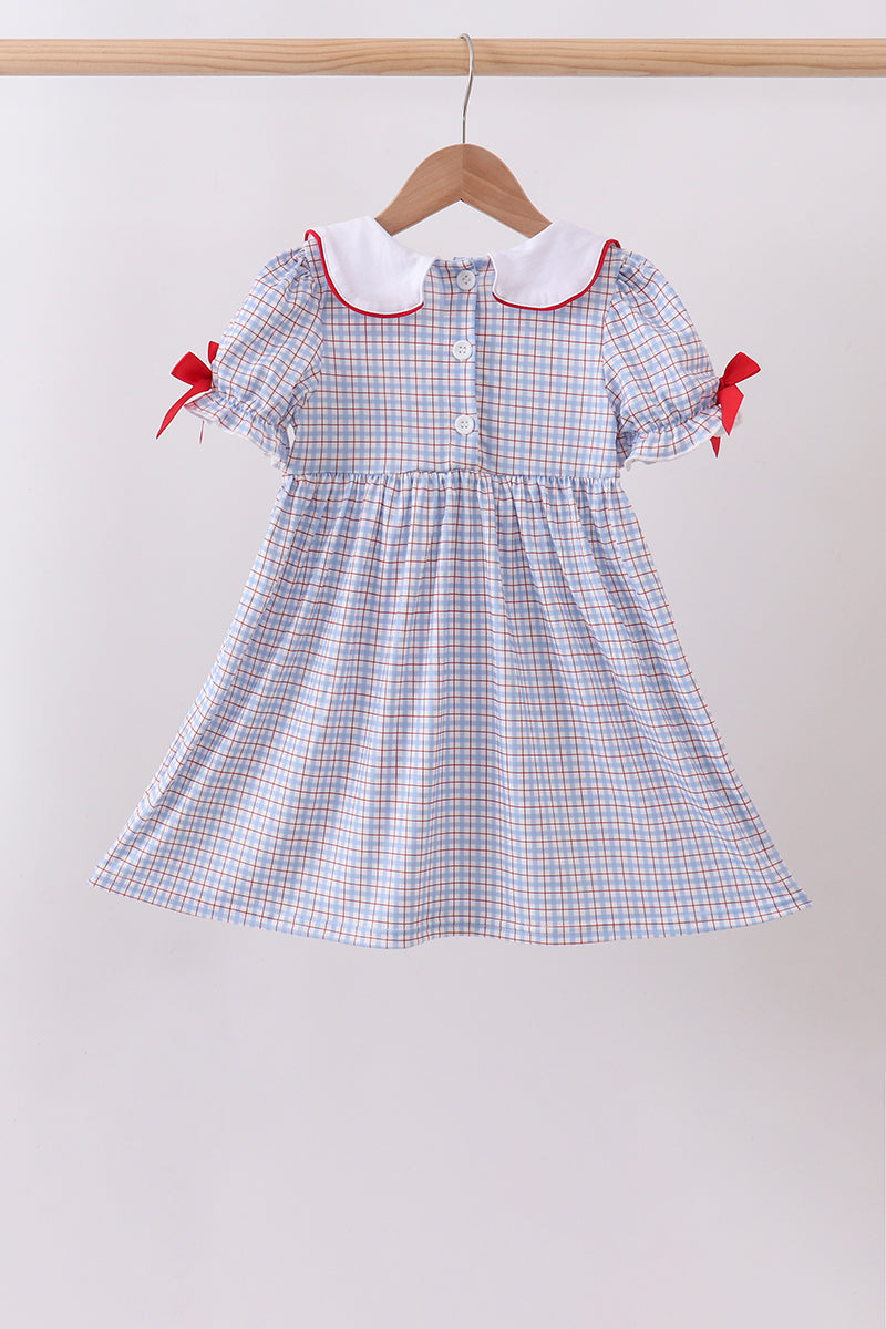 A toddler girl's short-sleeve dress with a light blue, white, and red gingham pattern, featuring a white Peter Pan collar with red trim and red ribbon bows on the sleeves, hanging on a wooden hanger.