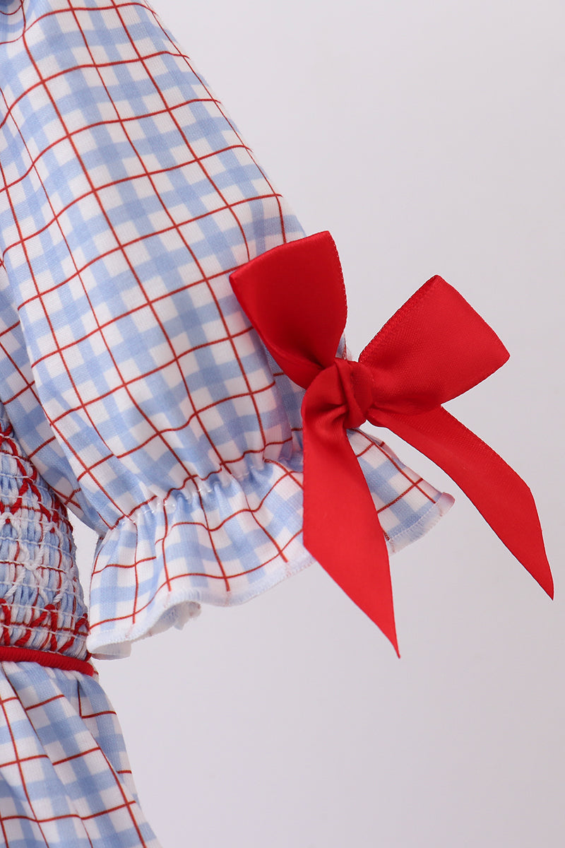 Close-up of a light blue and white gingham dress with red grid lines, featuring a smocked waist and a large red bow on the sleeve cuff.