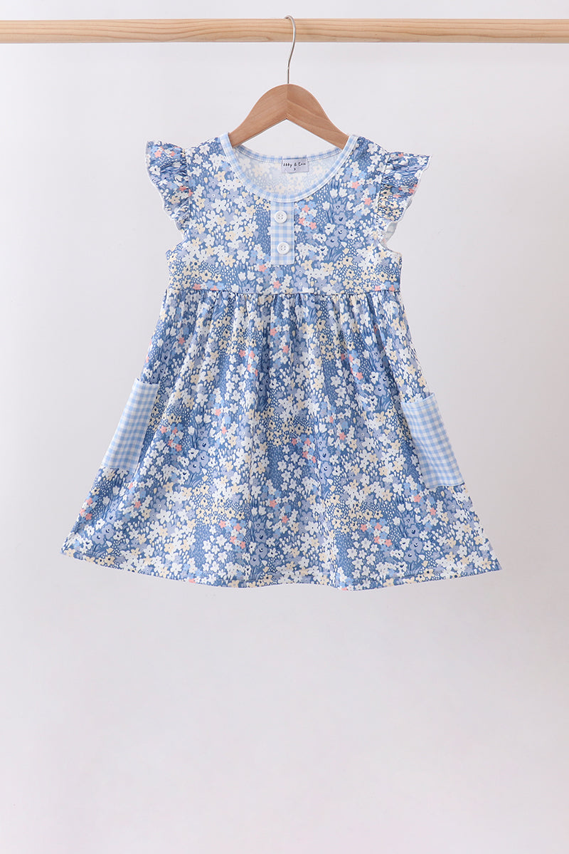 A blue and white floral print baby dress with flutter sleeves and gingham side pockets, hanging on a wooden hanger against a plain light background.