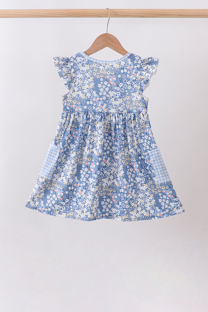 A blue and white floral print baby dress with ruffled sleeves and gingham side pockets, hanging on a wooden hanger against a plain light gray wall.