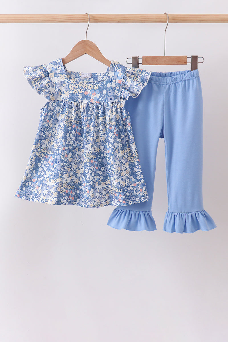 A toddler girl's two-piece clothing set hanging on wooden hangers against a plain light background. The set includes a light blue floral print smocked blouse with ruffled sleeves and matching light blue ruffled capri pants.