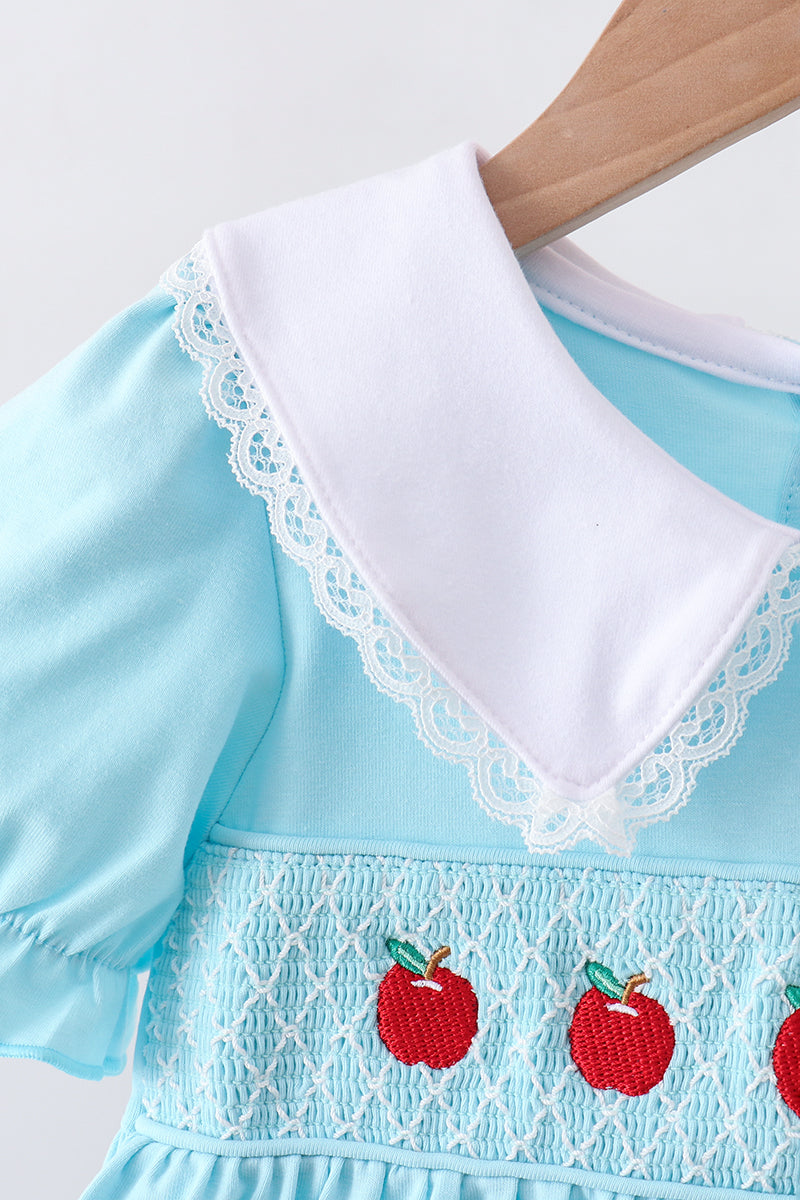 A close-up of a light blue baby dress with a large white collar trimmed in white lace, featuring embroidered red apples on the smocked front panel.