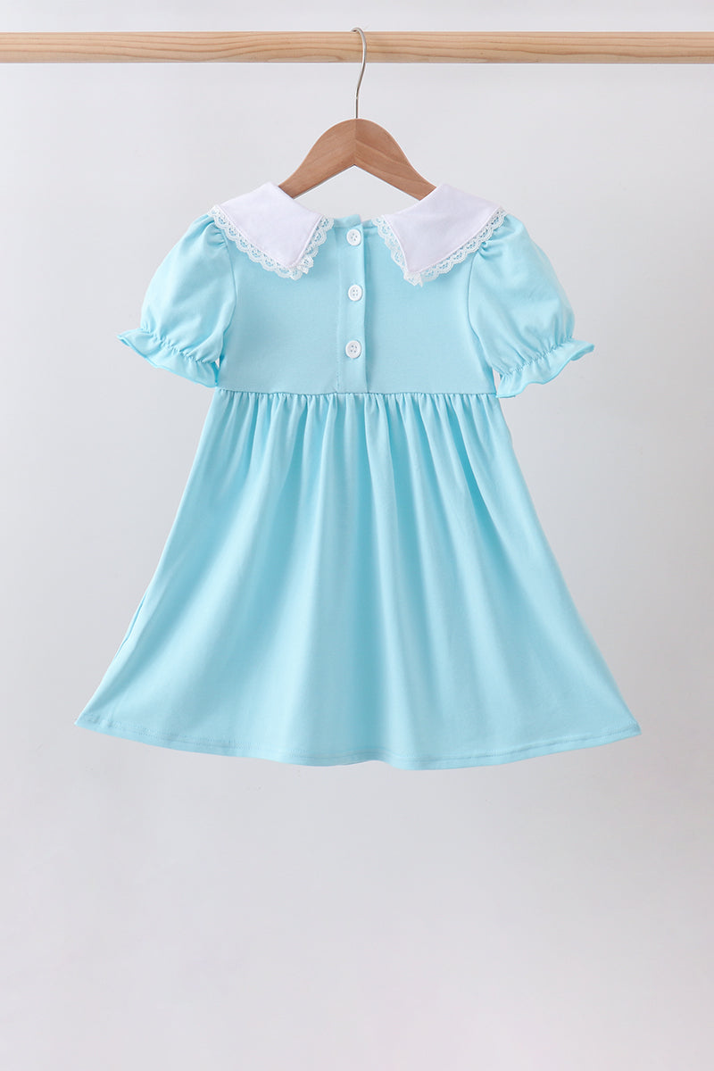 A light blue baby girl's dress with a white lace collar and button details, hanging on a wooden hanger against a plain light gray background.
