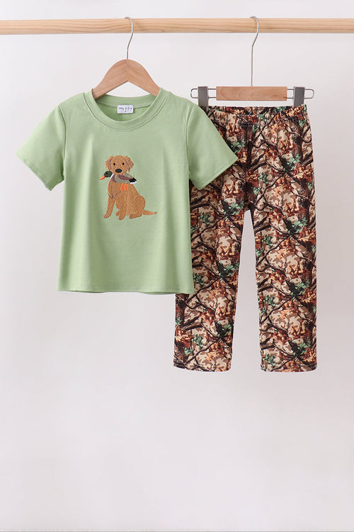 A two-piece children's outfit hanging on wooden hangers against a plain light background. The set includes a light sage green short-sleeve T-shirt with a brown dog graphic on the front, and matching camouflage-print pants in brown, green, and beige tones.