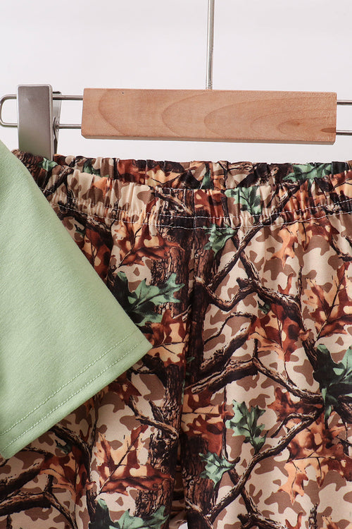 A close-up of a pair of camouflage-print shorts with an elastic waistband, hanging on a wooden hanger. A light green sleeve is draped over the left side of the shorts.
