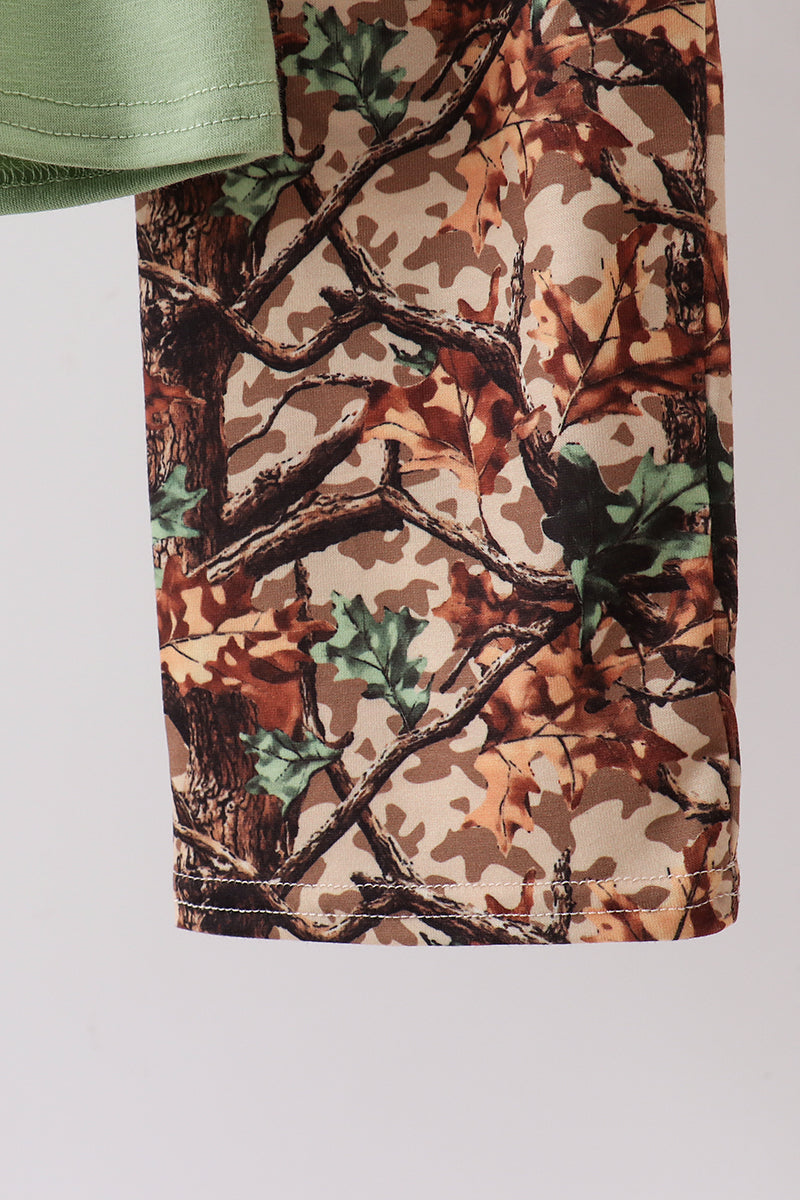 Close-up of camouflage shorts with a woodland tree-bark and leaf pattern in brown, beige, green, and black, with a light green shirt cuff visible at the top edge.