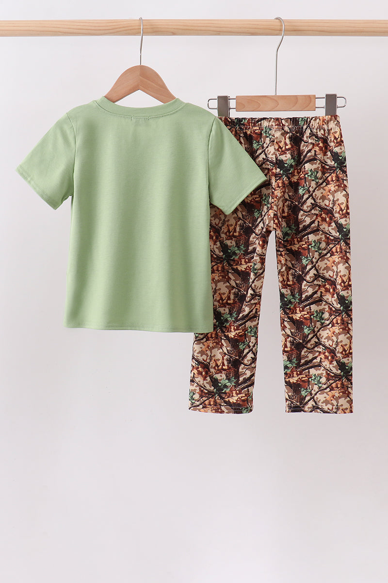 A children's two-piece outfit hanging on wooden hangers from a clothing rack. The set includes a light sage green short-sleeve t-shirt and matching camouflage-print pants in brown, green, and beige tones.