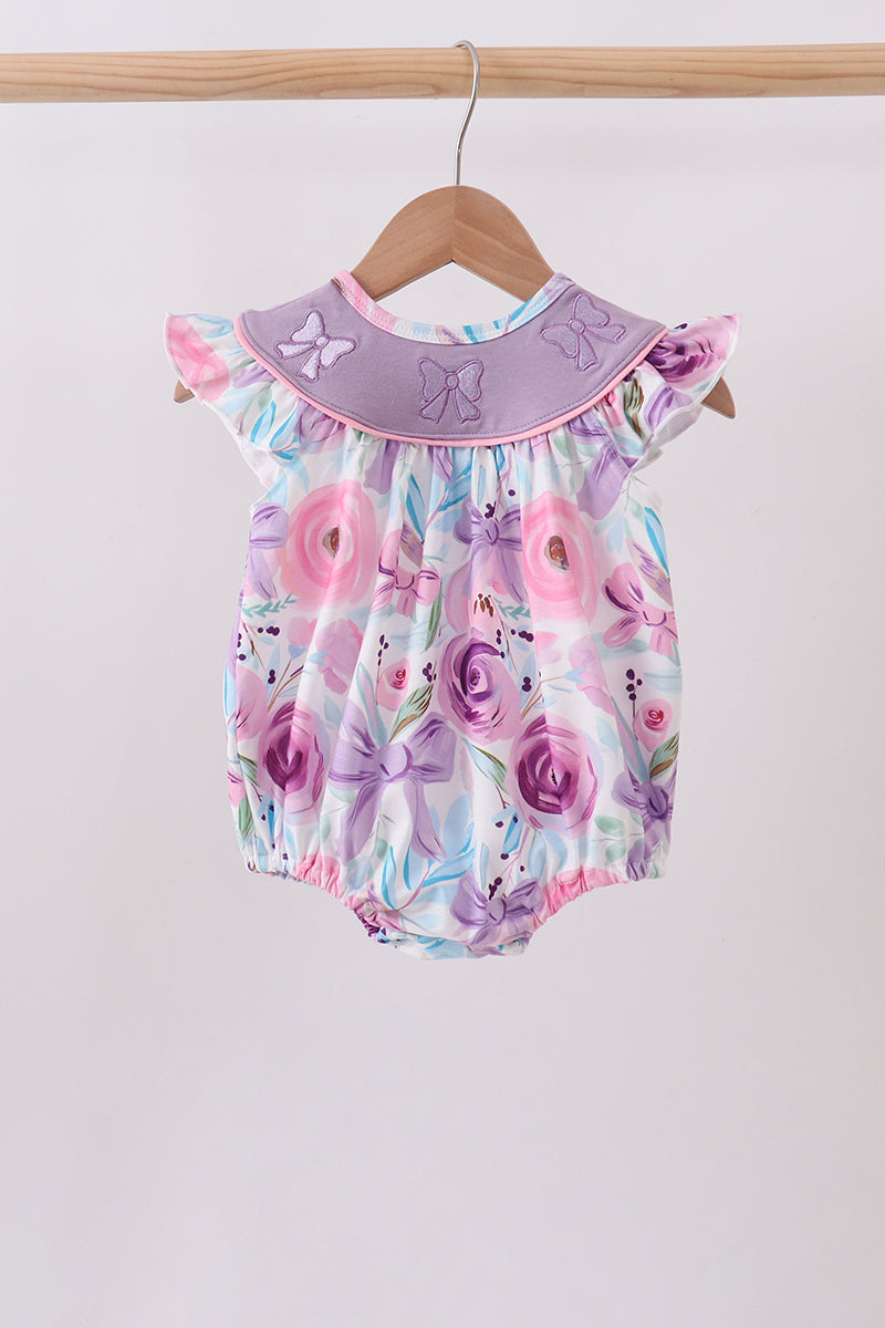 A baby romper with a purple and pink floral print, featuring a wide lavender collar with embroidered bows, hanging on a wooden hanger from a wooden clothes rack against a plain light gray wall.