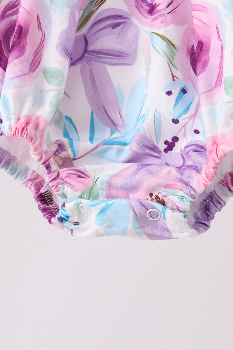 Close-up of a baby romper or bodysuit bottom with a pastel floral print in pink, purple, and light blue on a white background, featuring elastic ruffled trim and snap closures.