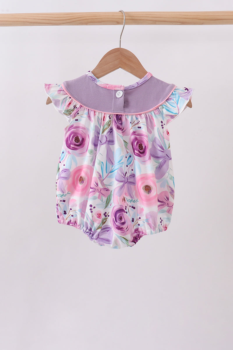A baby romper with a purple and pink floral print, featuring a solid lavender collar and ruffled sleeves, hanging on a wooden hanger against a plain light background.