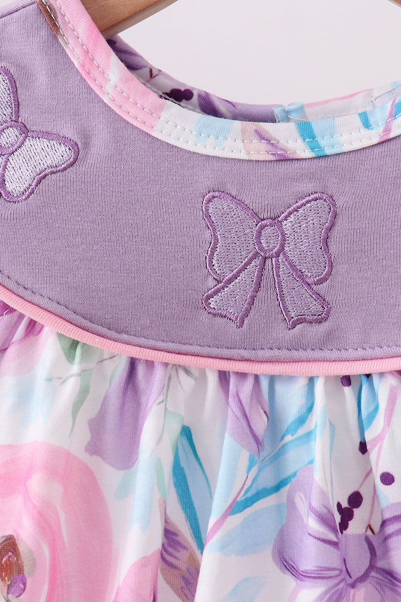 Close-up of a children's dress with a lavender purple collar featuring embroidered bow details, and a pastel floral skirt with pink, purple, and blue watercolor-style print.