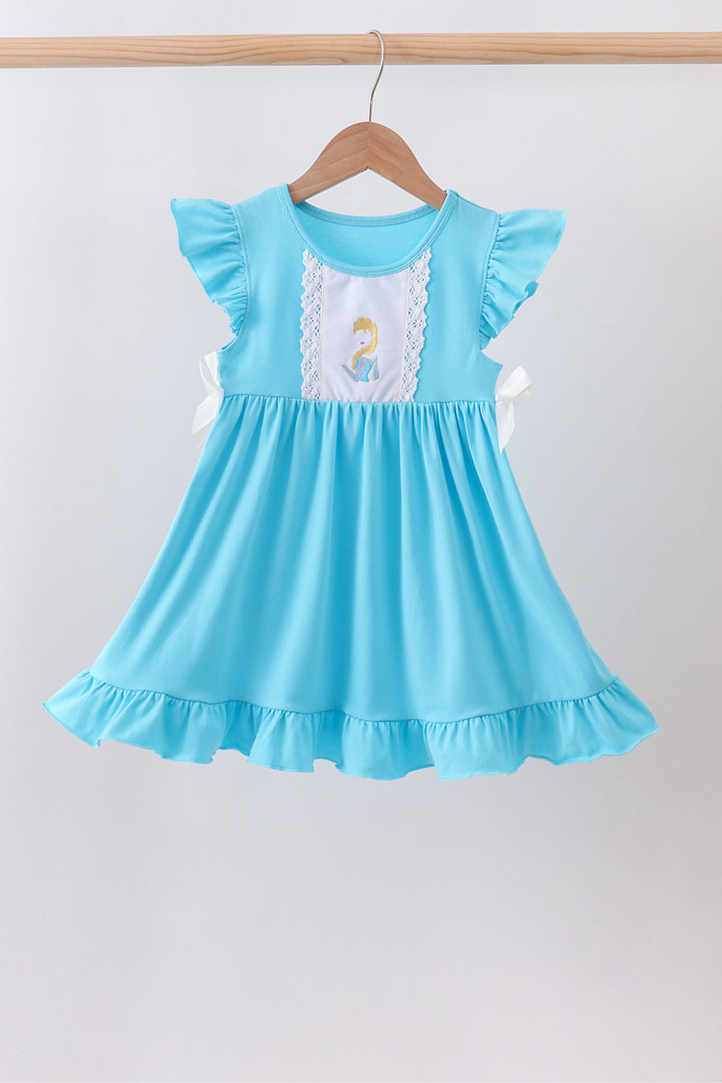 A light blue toddler girl's dress with ruffled cap sleeves, a white lace-trimmed chest panel, and a small embroidered character motif, hanging on a wooden hanger against a plain light gray wall.