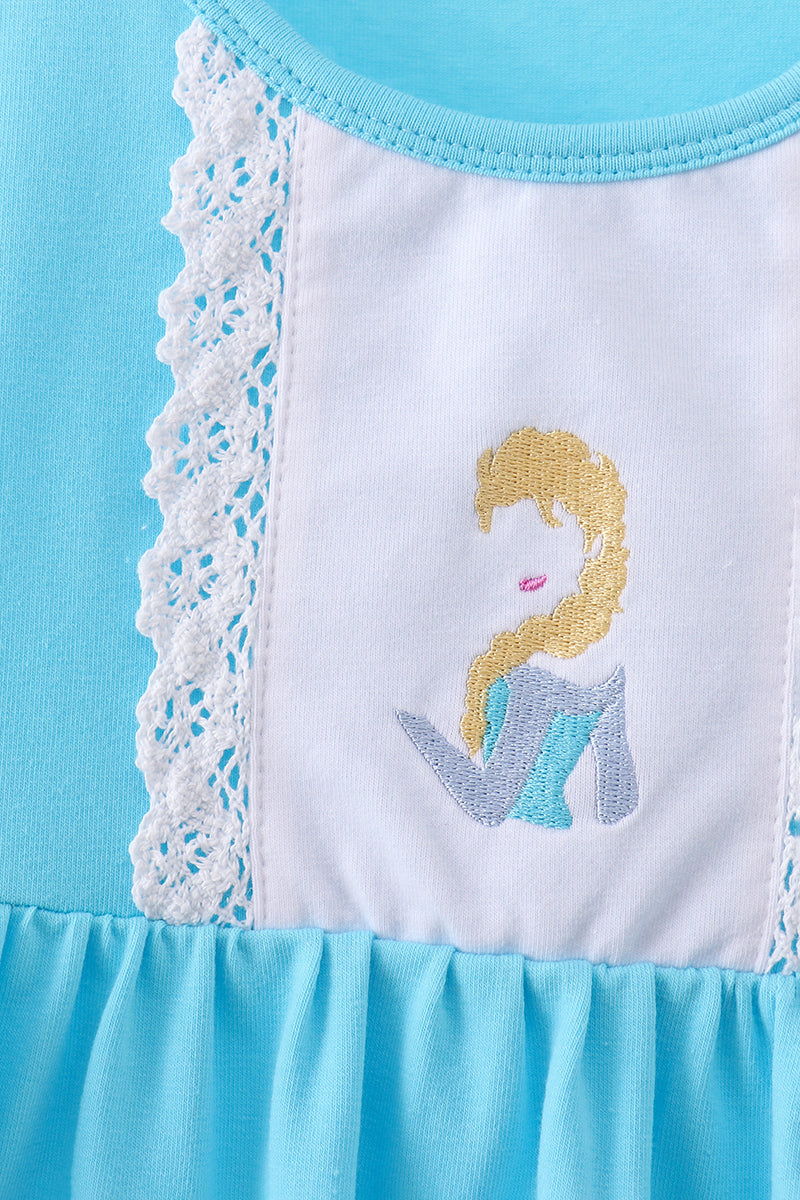 Close-up of a children's dress or top in light blue and white, featuring a central white panel with a stylized blonde character embroidery and white lace trim.