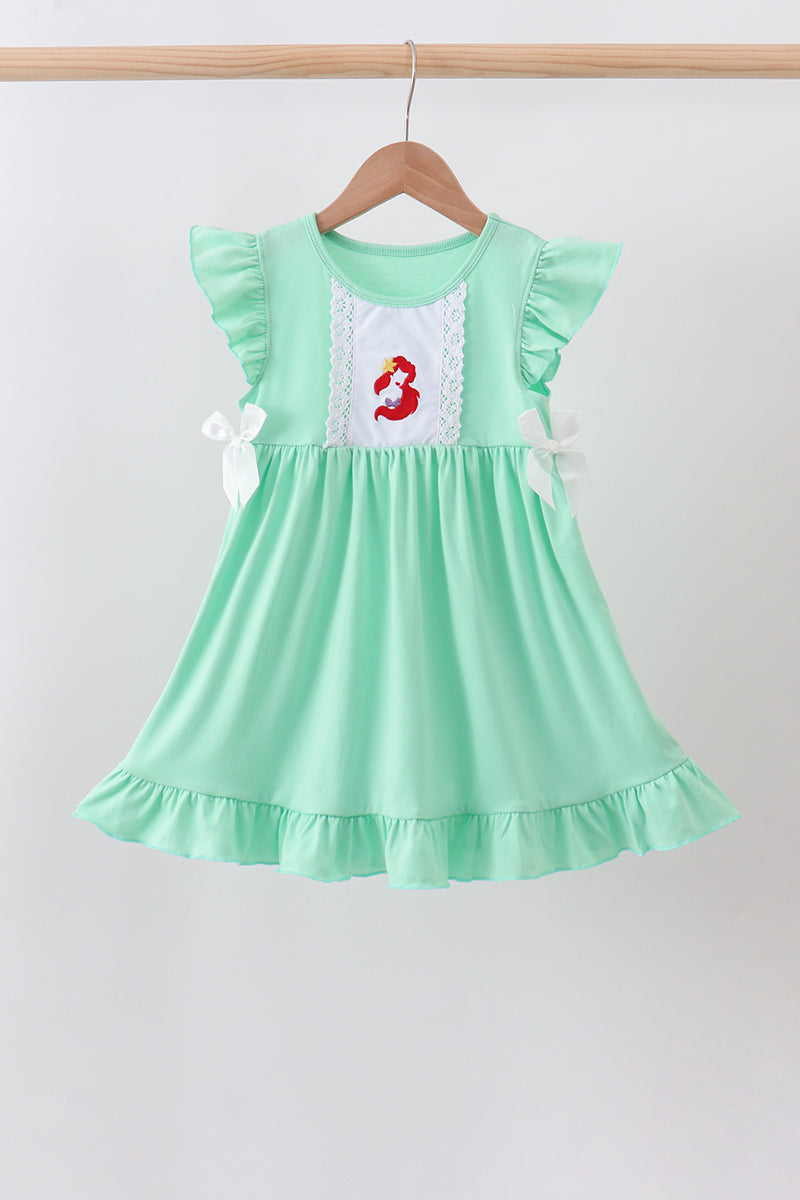 A mint green baby girl's dress hanging on a wooden hanger against a light gray background. The dress features ruffled sleeves, a gathered waist, a ruffled hem, and a white embroidered chest panel depicting the character Ariel from The Little Mermaid in red.