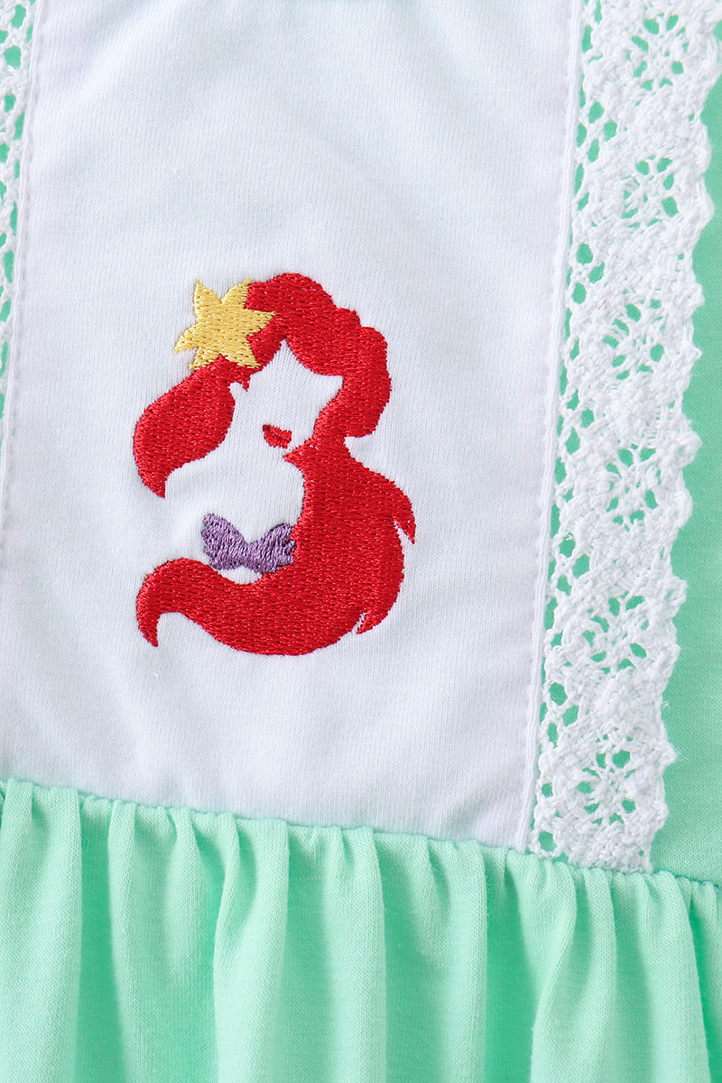 Close-up of a white and mint green garment featuring an embroidered mermaid design with red hair, a yellow star, and a purple bow, bordered by white lace trim.