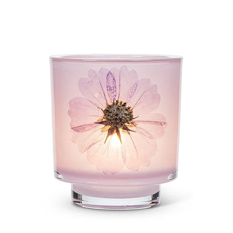 A pink glass candle jar with a pressed light purple flower embedded in the center, glowing with a warm light from within, set against a plain white background.