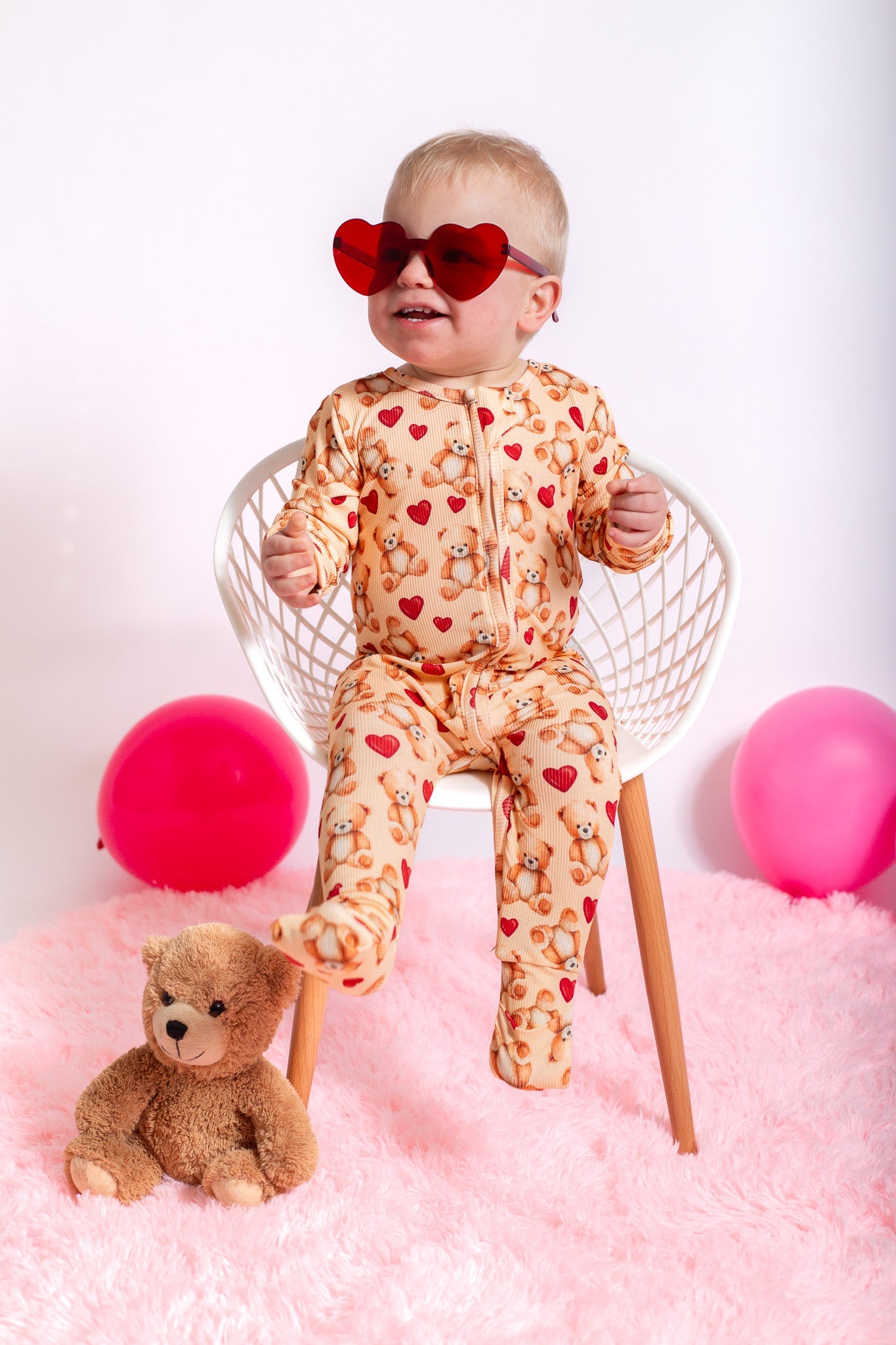 Baby in a patterned onesie with heart-shaped sunglasses, sitting on a chair with pink balloons and a teddy bear.