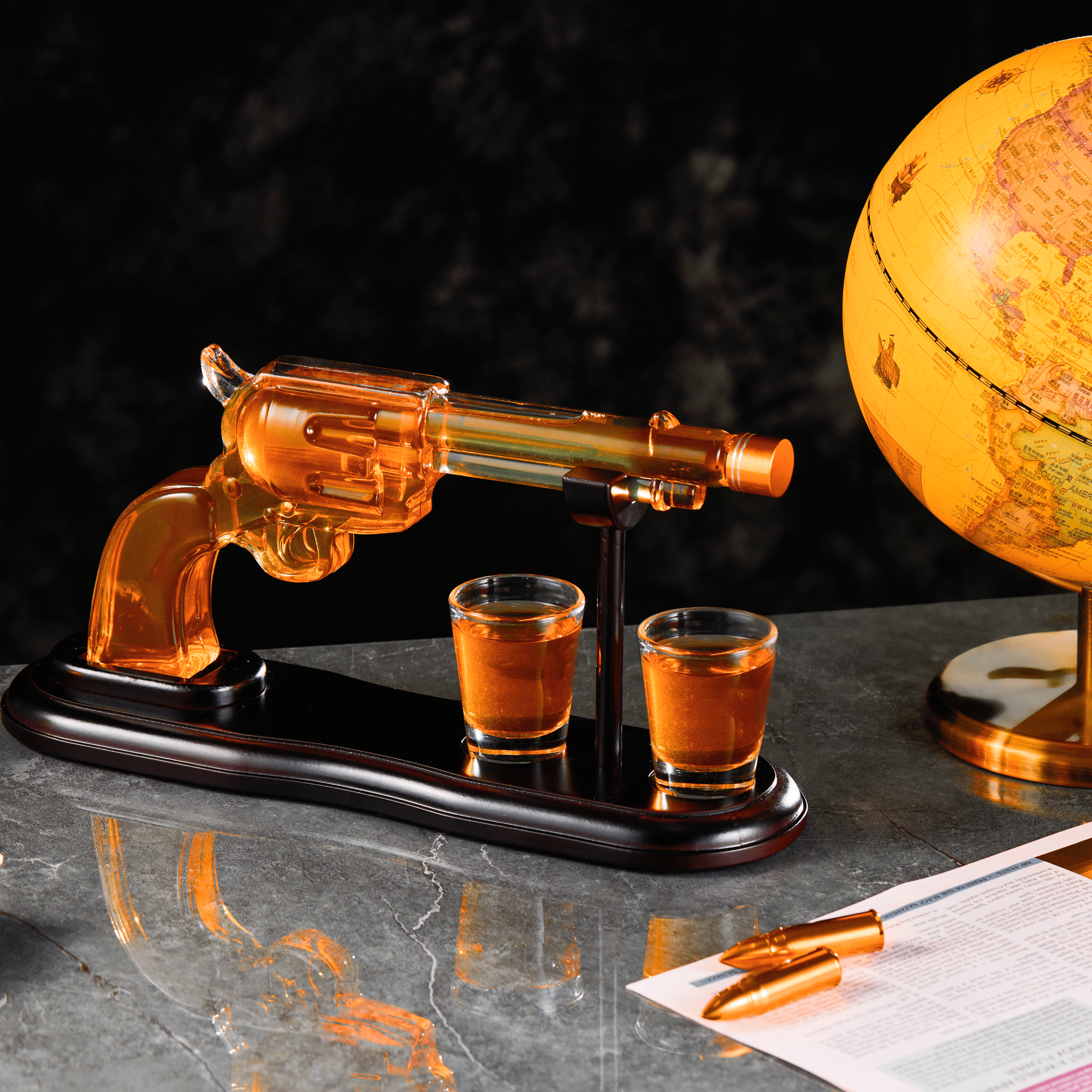 Glass revolver with two shot glasses on a stand next to a globe on a dark surface