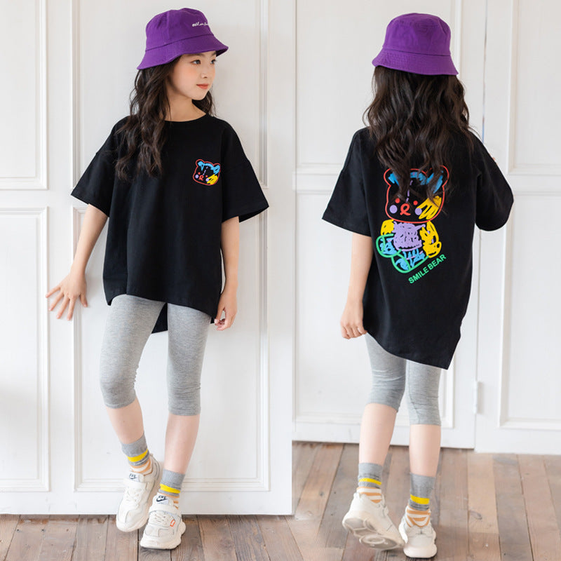 Two children wearing matching black t-shirts with colorful designs, gray leggings, and purple hats against a white paneled wall.