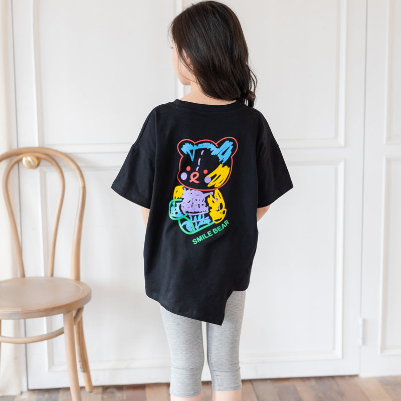 Person wearing a black t-shirt with a colorful bear graphic on the back, standing in a room with a wooden chair and white wall.
