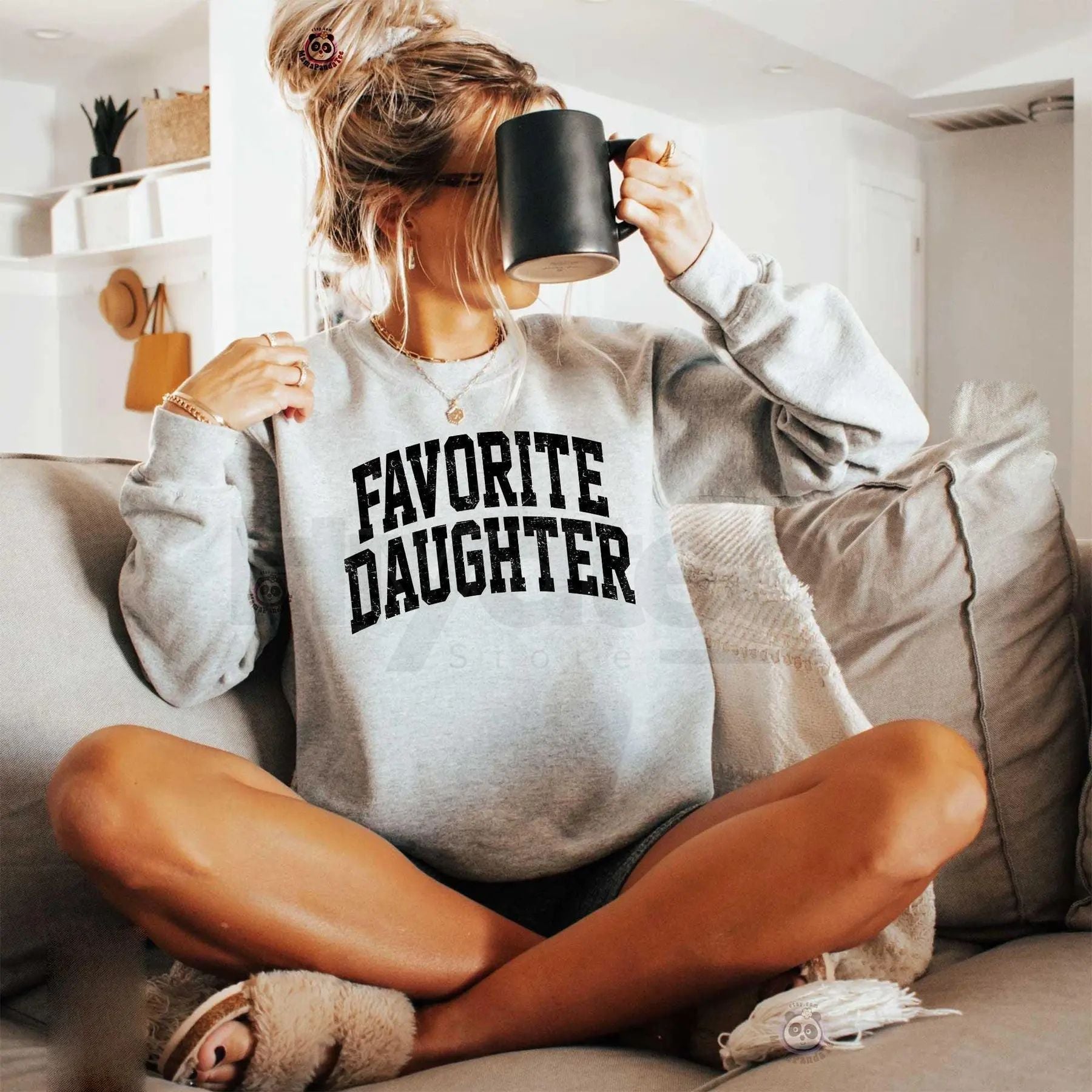 Woman wearing a 'Favorite Daughter' sweatshirt, sitting on a couch with a mug.