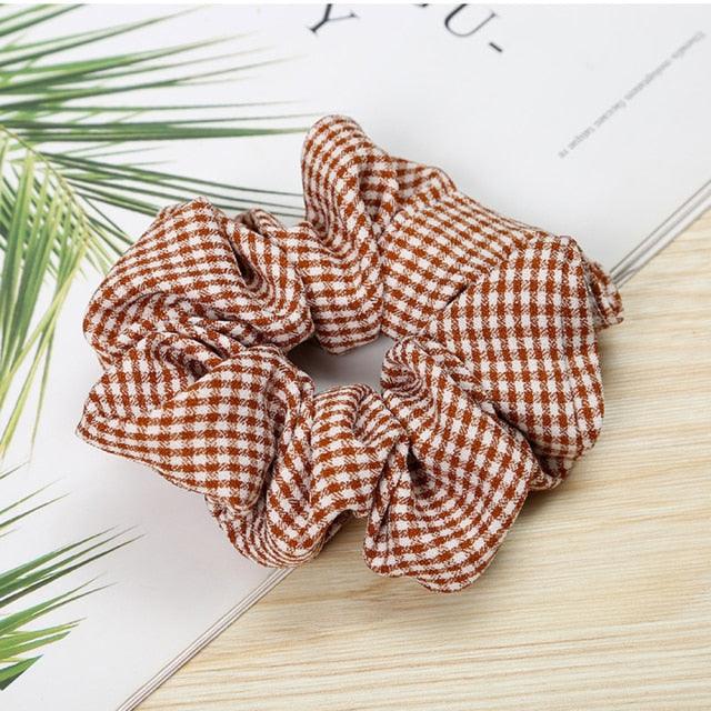 Three brown and white checkered scrunchies on a light wooden surface with green leaves in the corner.