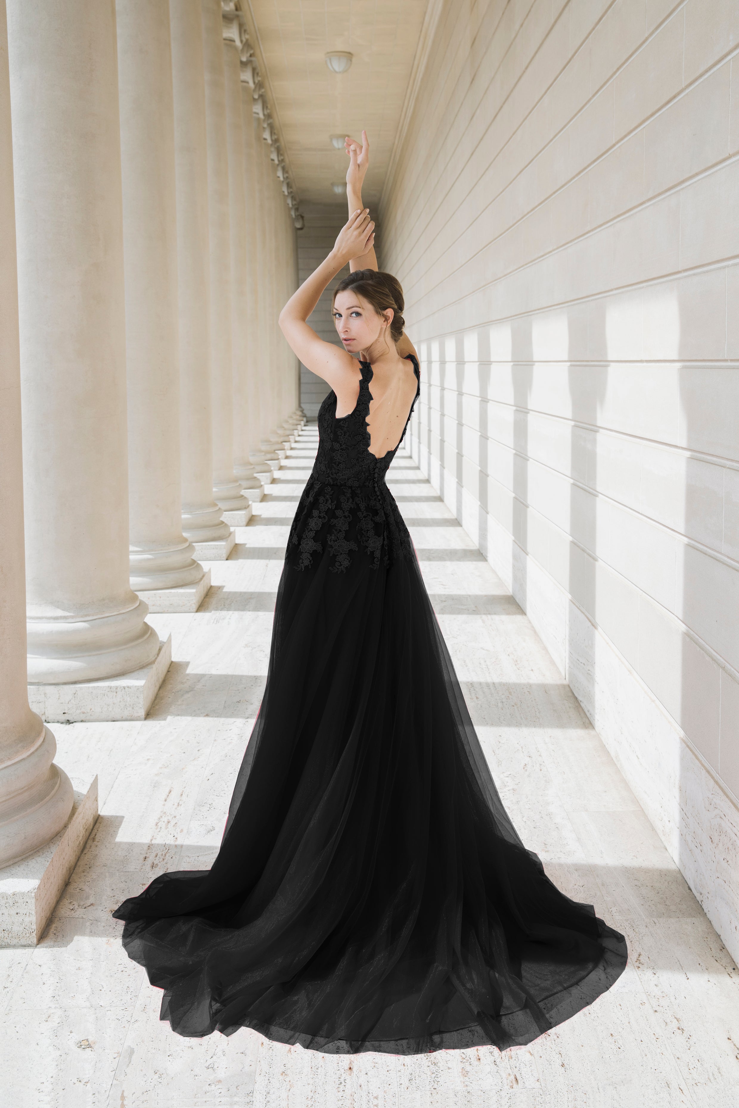 Woman in a black evening gown standing in a sunlit colonnade.
