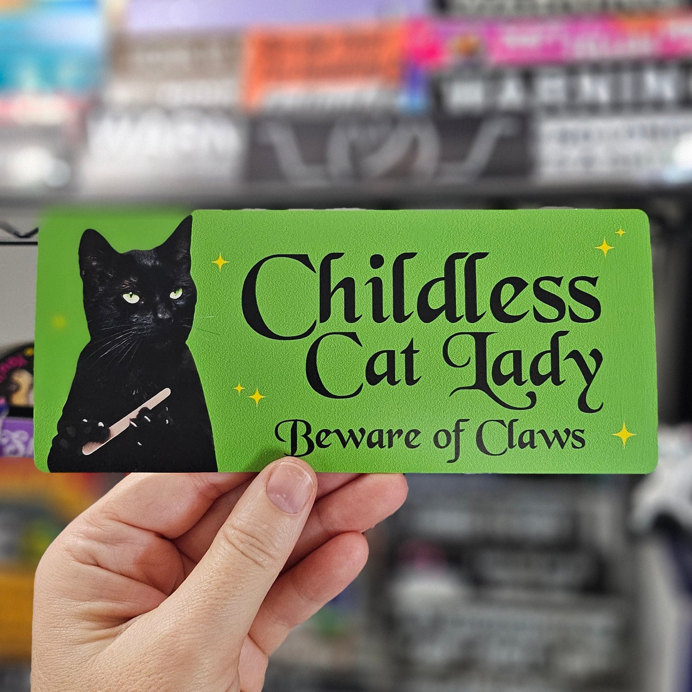 Card with a black cat illustration and text 'Childless Cat Lady Beware of Claws' held by a hand in front of a blurred store background.