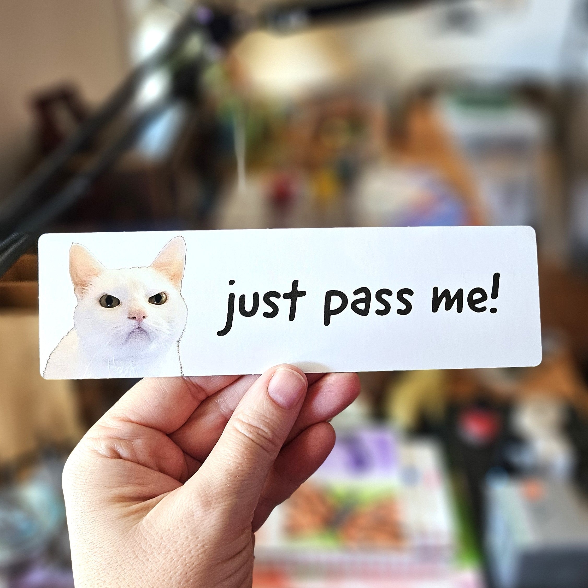 Hand holding a card with a cat illustration and text 'just pass me!' in a blurred indoor setting.