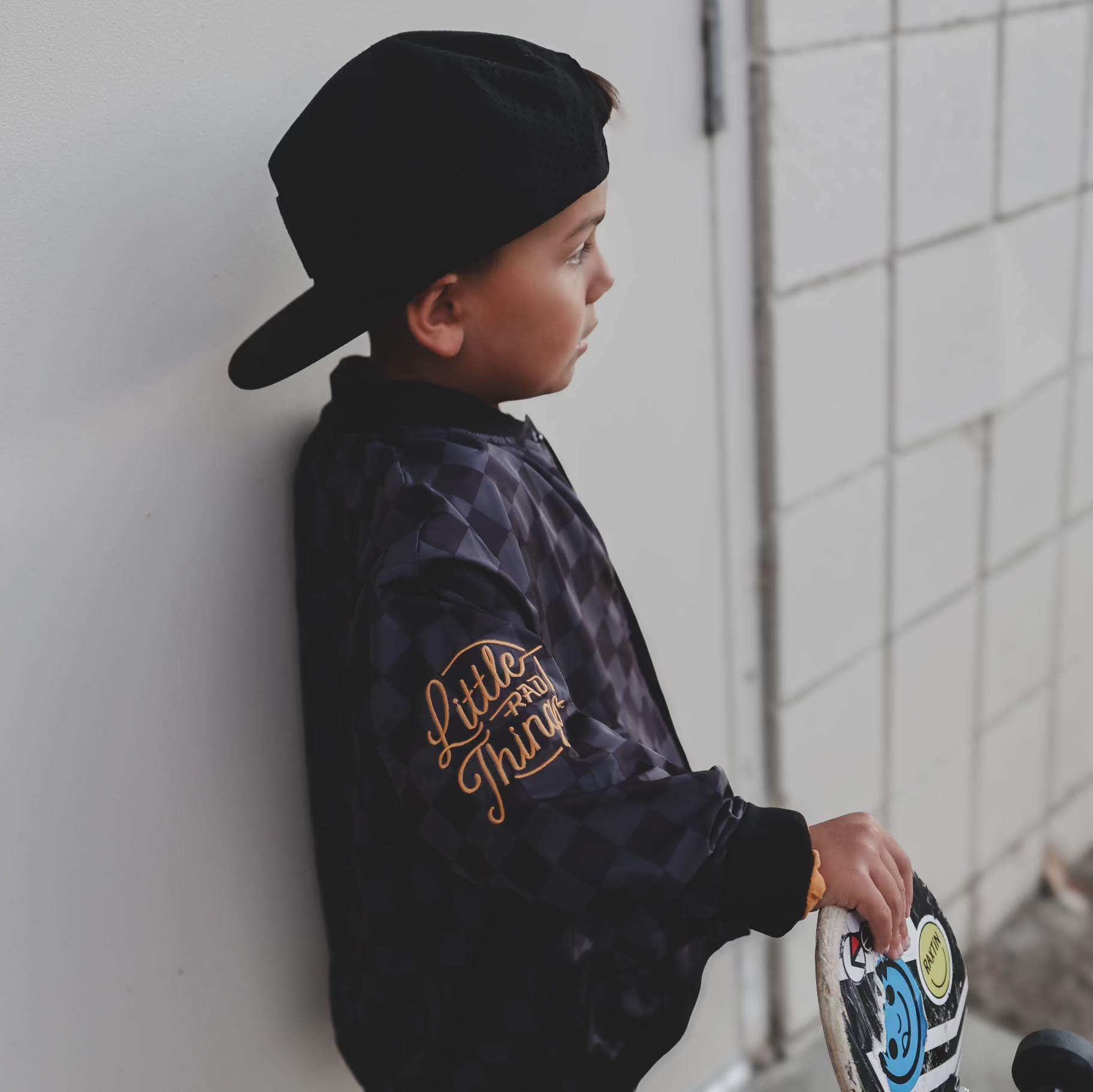 Child wearing a black jacket with 'Little Thing' text, leaning against a wall.