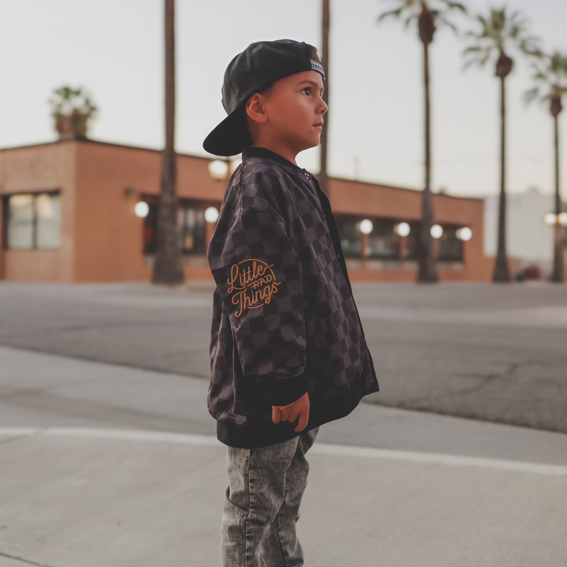 Child wearing a jacket with 'Little Things' text in an urban setting with palm trees.