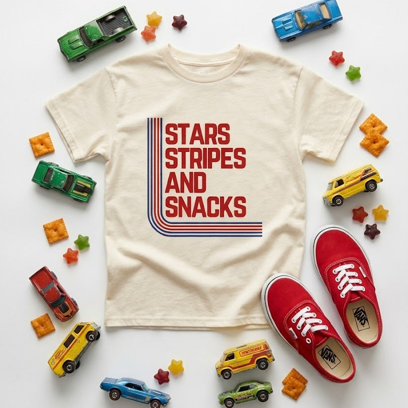 Beige t-shirt with 'Stars Stripes and Snacks' design, surrounded by toy cars and snacks on a white background