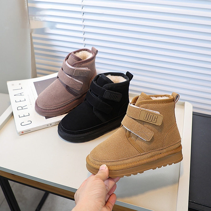 Three pairs of children's boots in brown, black, and beige on a table with a book underneath.