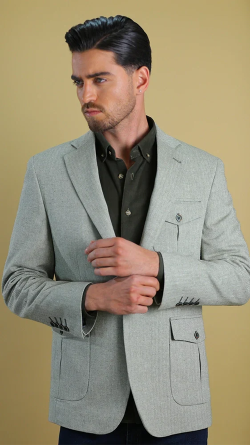 A man wearing a light gray textured blazer over a dark olive green button-down shirt, posed against a solid warm beige background.