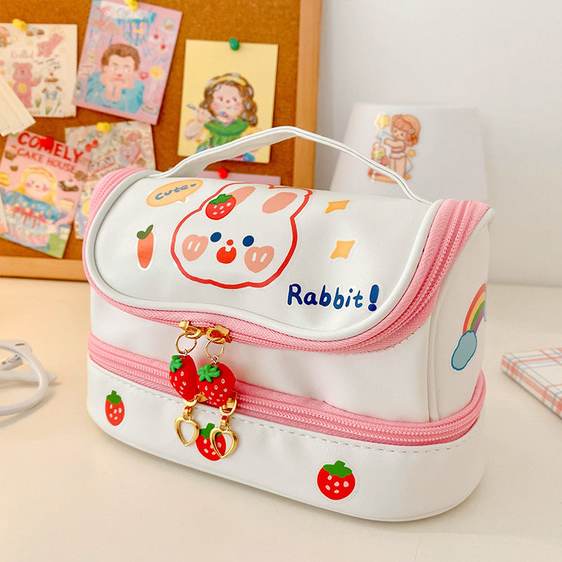 A white pencil case with pink trim, featuring a cartoon rabbit face, strawberries, a rainbow, and the word 'Rabbit!' on the front, shown on a desk with stationery in the background.