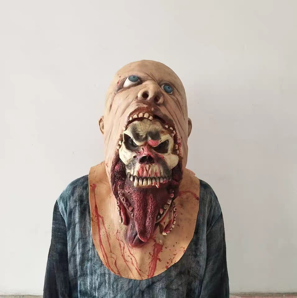 A horror Halloween mask with a grotesque stitched mouth, exposed skull, bloody details, and blue eyes, worn by a person in a dark shirt against a plain light wall.