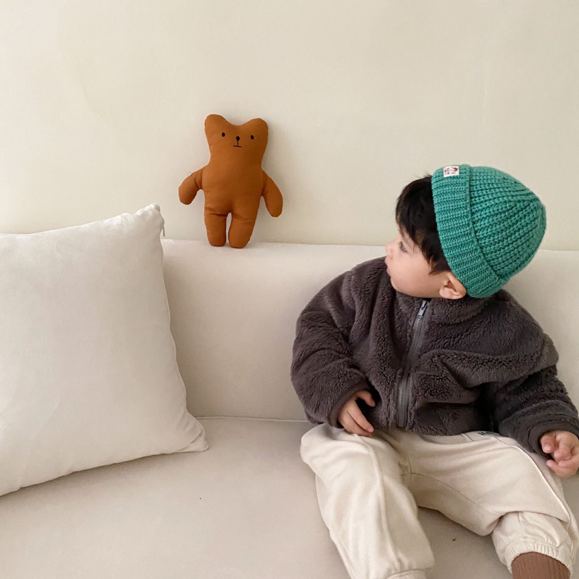 Child sitting on a couch wearing a green beanie with a teddy bear toy beside them.