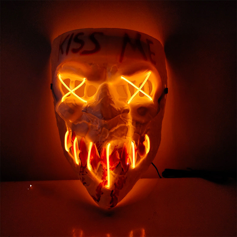 A skull-shaped LED light-up mask with glowing orange eyes and teeth, featuring the words 'KISS ME' written on the forehead, set against a dark background.