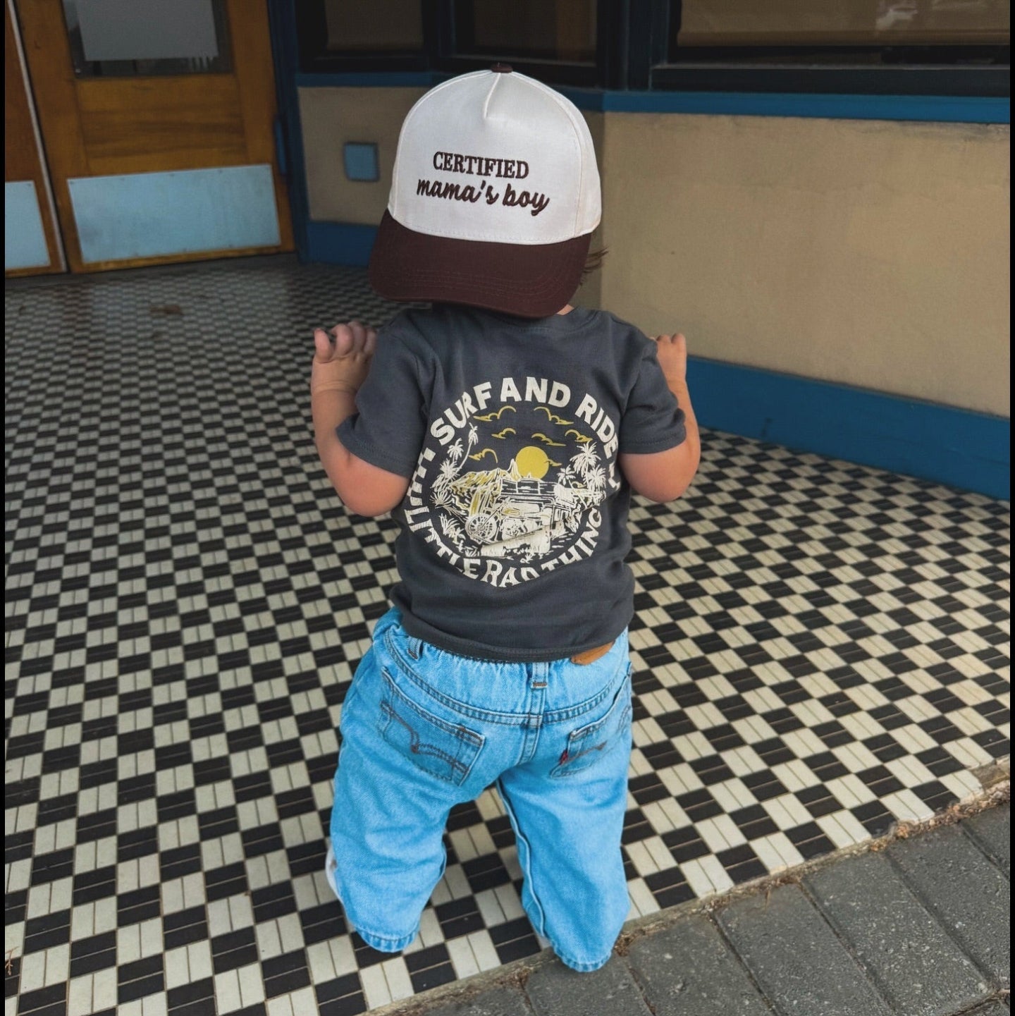 Child wearing a cap and t-shirt with text, standing on a checkered floor.