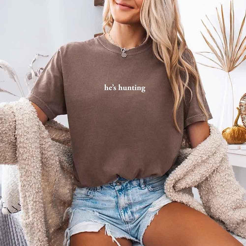 Person wearing a brown t-shirt with 'he's hunting' text, sitting indoors.