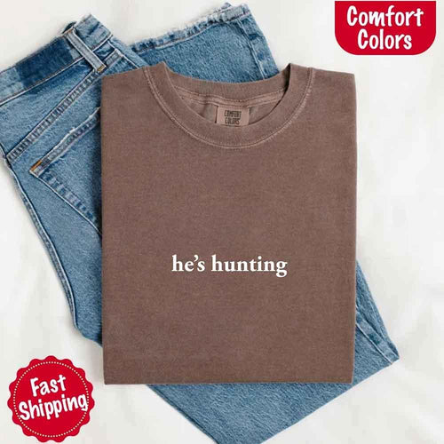 Folded brown t-shirt with 'he's hunting' text on blue jeans, branded 'Comfort Colors'.